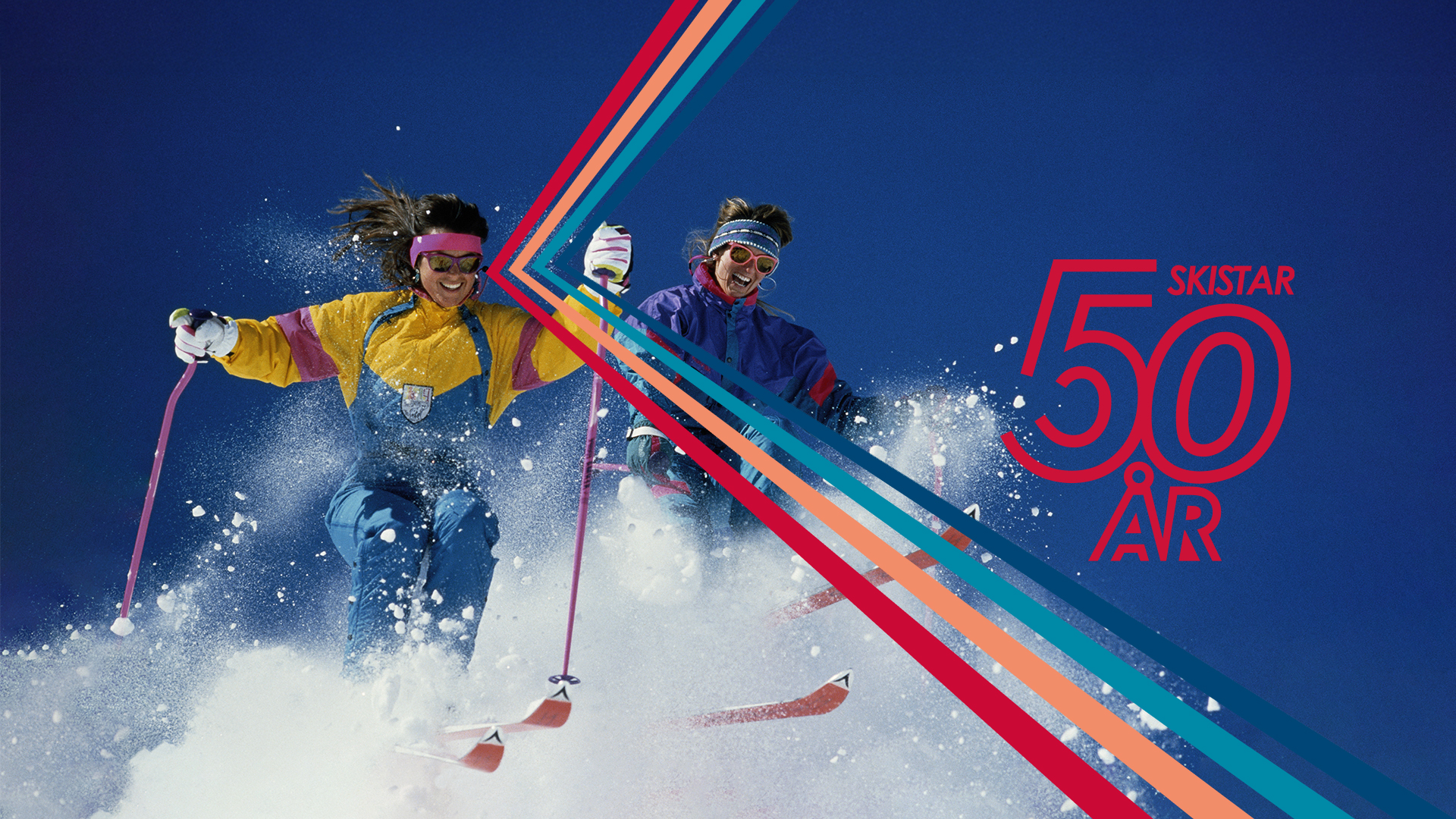 Två personer åker skidor nerför en snöig backe under klarblå himmel, med färgglada skidkläder och skidglasögon. Texten "Skistar 50 år" syns på högra sidan av bilden