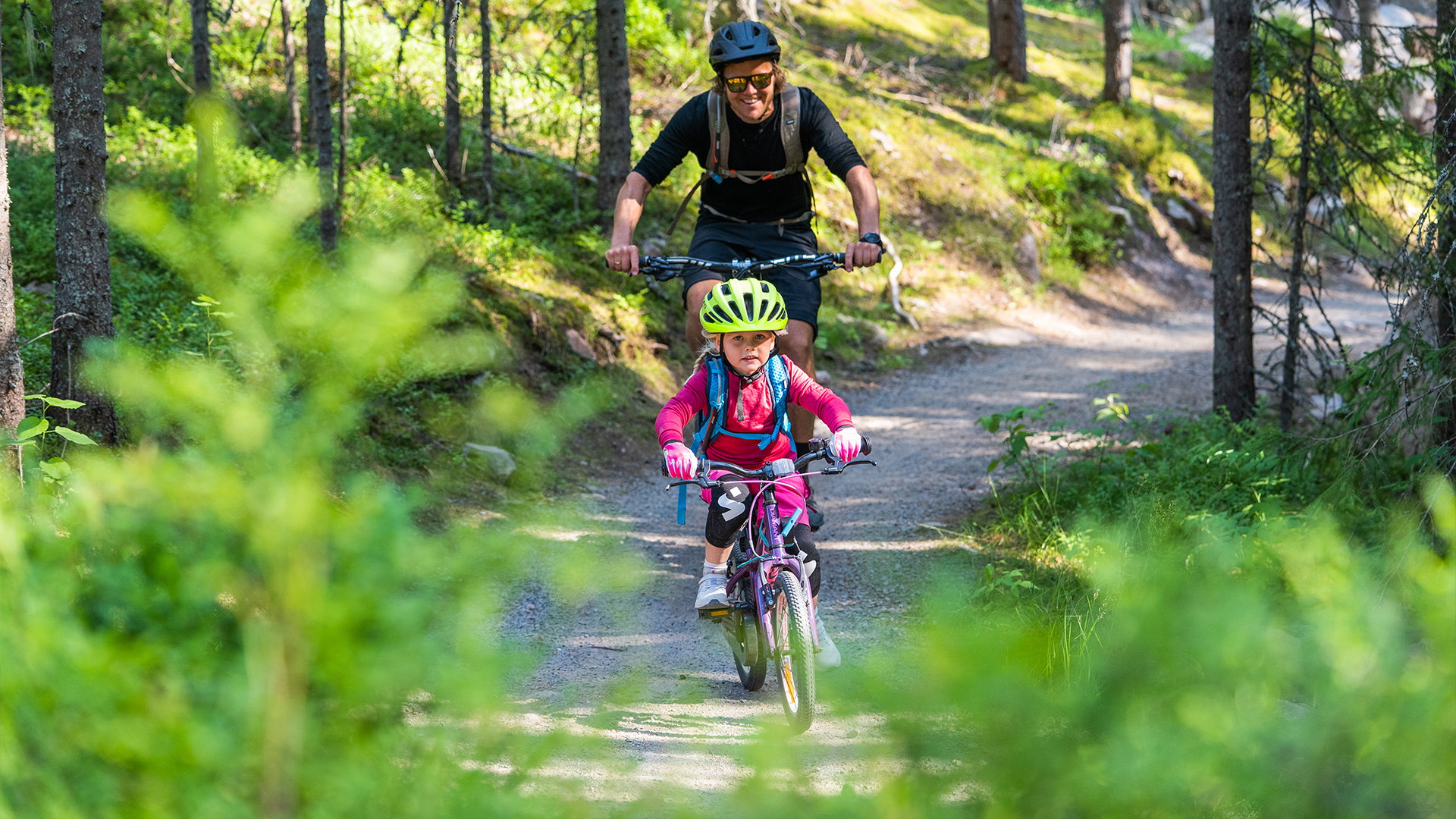 Person cyklar med barn genom skogstig på solig dag, iklädda hjälmar och cykelkläder