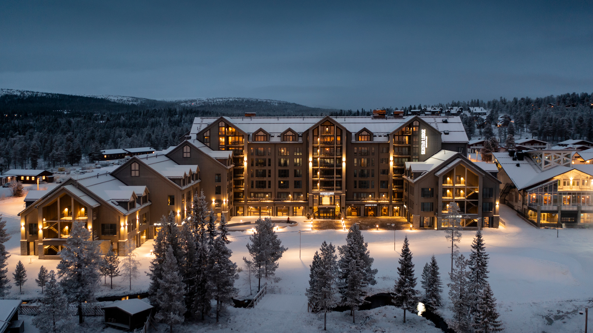 Hotel in a winter landscape with snow-covered ground and buildings lit up at dusk, surrounded by frosty trees and mountains